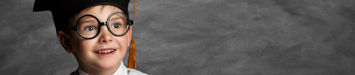 Enfant souriant portant un chapeau de diplômé et des lunettes, bras croisés, devant un tableau noir.