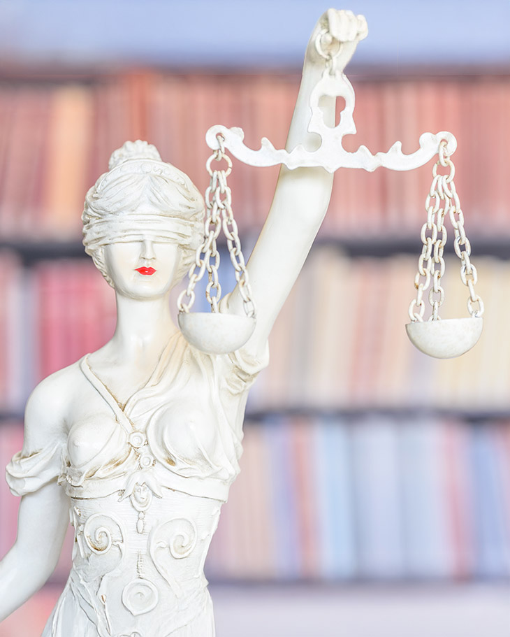 Statue de la Justice tenant une balance, les yeux bandés, devant une bibliothèque de livres juridiques.